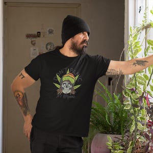 Könnte beinhalten: Schwarzes T-Shirt mit einem Totenkopf, der einen Rasta-Hut trägt, und dem Text "DON'T PANIC It's Organic". Das T-Shirt wird von einer Person mit Tätowierungen und einer schwarzen Mütze getragen.
