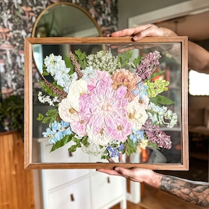 May include: A framed pressed flower arrangement with a variety of flowers in shades of pink, white, blue, and purple. The flowers are arranged in a bouquet shape and are pressed between two pieces of glass. The frame is made of wood and has a natural finish.