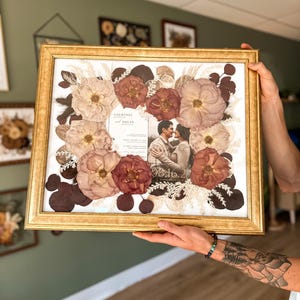 May include: A gold-framed picture with a pressed flower arrangement and a wedding invitation inside. The invitation reads "Courtney & Nolan" and the date "08.16.22".