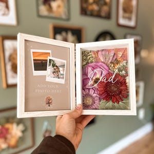 May include: A white, hinged picture frame with two sections. One side displays two photos, the other features dried flowers with the word "Dad" in script. The frame is held against a backdrop of framed art.