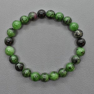 Könnte beinhalten: Ein Armband mit runden Perlen in Grün und Schwarz. Die Perlen sind poliert und weisen eine Mischung aus einfarbigem Grün und gesprenkeltem Schwarz auf. Das Armband wird auf einer grauen Oberfläche präsentiert. Die Perlen haben einen Durchmesser von ca. 1 cm.