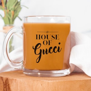 May include: A clear glass mug with a brown liquid inside. The mug has a black design that says "HOUSE OF GUCCI" on it.