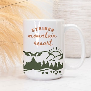 May include: White ceramic mug with a brown and green mountain scene and the text "Steiner mountain resort" in brown.