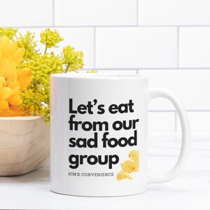 May include: White ceramic mug with black text that reads "Let's eat from our sad food group" and a small image of potato chips. The text "Kim's Convenience" is printed in smaller font below the main text.