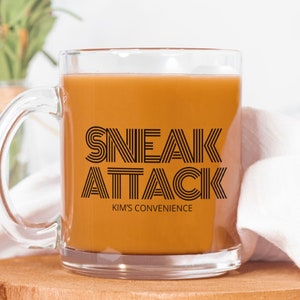 May include: A clear glass mug filled with a light brown liquid, possibly coffee or tea. The mug features the words "SNEAK ATTACK" in a bold, black, retro font, with "KIM'S CONVENIENCE" below. The mug sits on a wooden surface.