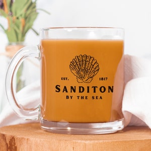 May include: A clear glass mug with a brown beverage inside. The mug has a black logo that says "EST. 1817 SANDITON BY THE SEA" with a seashell graphic.