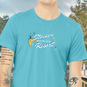 May include: A turquoise blue t-shirt with a retro-style logo that says "Steiner Mountain Resort" in white and blue lettering. The logo features a stylized mountain with a starburst design.