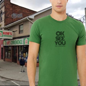 May include: Green t-shirt with black text that reads "OK SEE YOU" and "KIM'S CONVENIENCE" underneath.