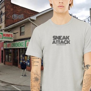 May include: A light gray t-shirt with a black graphic that says "SNEAK ATTACK" and "KIM'S CONVENIENCE" in a retro font.
