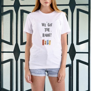 May include: White t-shirt with the text "We Got The Rabbi!" in black lettering. The text is surrounded by small stars. There are five colorful rectangles below the text.