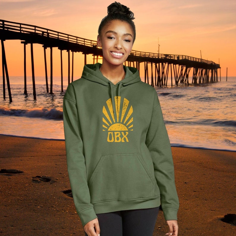 May include: A green hooded sweatshirt with a yellow sun and "OBX" text on the front. The sweatshirt is worn by a person standing on a beach with a pier in the background.