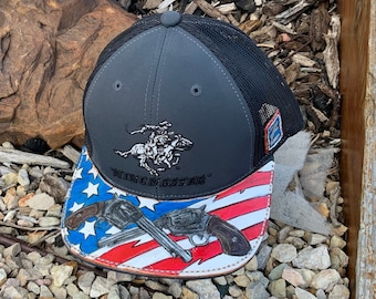 May include: A gray and black baseball cap with a mesh back. The front features a cowboy on a horse and the word "WINCHESTER". The brim is decorated with an American flag design and two crossed revolvers.