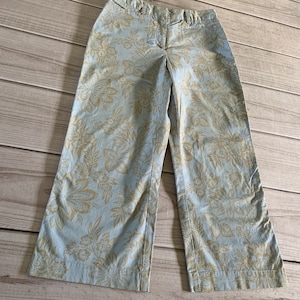 May include: Light blue wide-leg pants with a beige floral pattern. The pants have a button and zipper closure and two front pockets. The fabric appears to be cotton or a cotton blend. The pants are laid flat on a wooden surface.