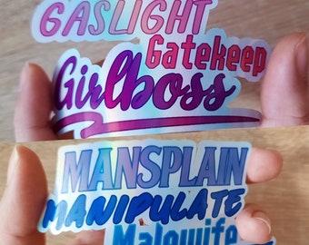 May include: Two iridescent stickers with text. The top sticker is pink, blue, and purple with the text "Gaslight Gatekeep Girlboss". The bottom sticker is blue and purple with the text "Mansplain Manipulate Malewife".