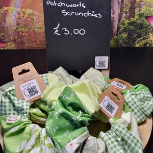 May include: A selection of green patchwork scrunchies with floral and gingham patterns. The scrunchies are displayed on a wooden surface, with a chalkboard sign that reads "THE GREEN ONE... Patchwork Scrunchies."