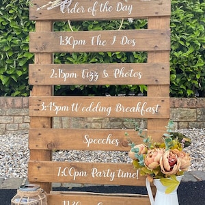 May include: A wooden wedding sign displaying the day's schedule, including "The I Dos", "Fizz & Photos", and "Party time!". The sign is adorned with flowers and a decorative jar. The sign's dimensions are not visible.