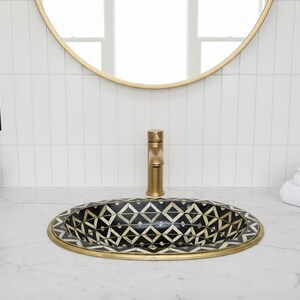 Può includere: Un lavabo ovale con un motivo geometrico bianco e nero, incorniciato da un bordo dorato. Un rubinetto dorato è centrato sopra il lavabo. Il lavabo è posto su un piano di lavoro bianco, con uno specchio con cornice dorata sopra.