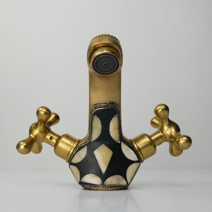 May include: A brass tap with a decorative design. The tap has two gold-coloured handles and a central body with a black and white inlaid pattern. The spout has a mesh screen.