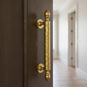 Könnte beinhalten: Nahaufnahme einer dunkelbraunen Holztür mit einem dekorativen goldfarbenen Griff. Der Griff hat ein verziertes Design und ist mit kreisförmigen Platten an der Tür befestigt. Der Hintergrund zeigt einen Flur.