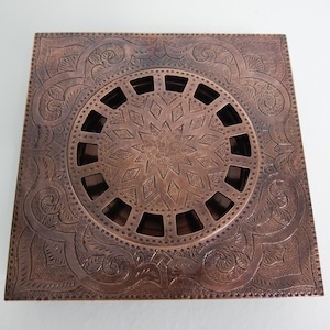 Könnte beinhalten: Quadratischer Kupfer-Abdeckrost mit aufwendigen floralen und geometrischen Mustern. Die Mitte zeigt ein kreisförmiges Design mit einem Sternmuster und rechteckigen Öffnungen. Die Ränder sind mit detaillierten Ranken und floralen Motiven verziert.