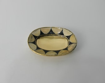 May include: Oval decorative tray with a gold interior and a patterned border of black and cream-colored shapes. The tray has a gold-colored rim and a textured edge.