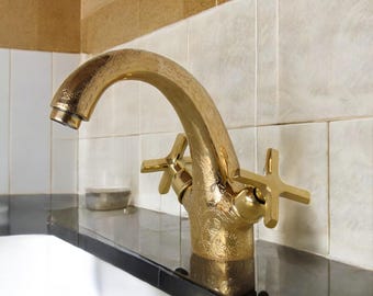 Op de afbeelding: Een gouden badkamerkraan met een sierlijk ontwerp. De kraan heeft twee kruisvormige handvatten en een gebogen uitloop.