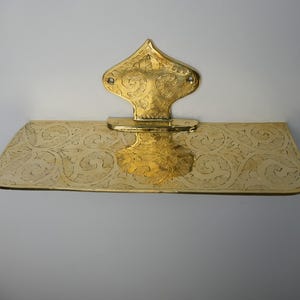 May include: A gold-colored, ornate wall-mounted shelf with a decorative backplate. The shelf and backplate feature intricate etched patterns. The backplate has a stylized, pointed top. The shelf is rectangular with rounded corners.