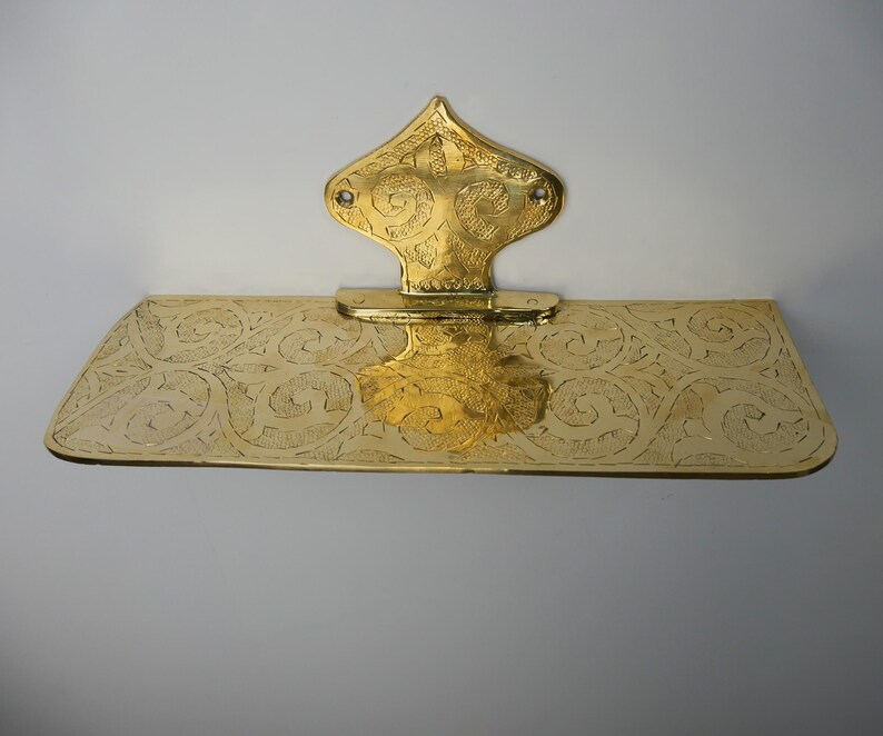 May include: A gold-colored, ornate wall-mounted shelf with a decorative backplate. The shelf and backplate feature intricate etched patterns. The backplate has a stylized, pointed top. The shelf is rectangular with rounded corners.
