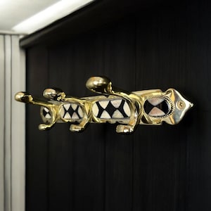 May include: A brass coat rack with three hooks and a black and white chequered design. The rack is mounted on a dark wood wall.