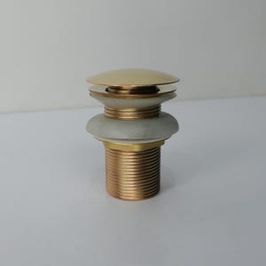May include: A gold-toned pop-up drain stopper with a threaded base. The stopper has a rounded top and a white and gold midsection. The drain stopper is designed for use in a bathroom sink.