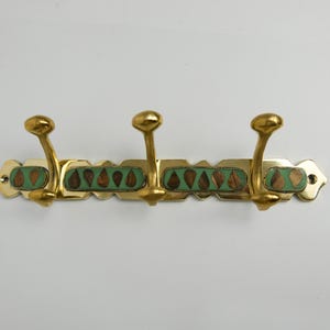May include: A brass wall-mounted coat rack with three curved hooks. The rack has decorative green and brown inlaid accents. The hooks and the rack have a shiny, golden finish. The rack is designed to hold coats, hats, or other items.