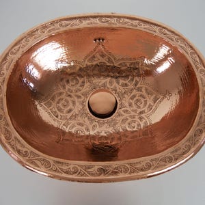 May include: Oval copper sink with an etched floral pattern. The sink has a hammered texture and a decorative border. The interior features a central drain and a detailed pattern. The copper finish gives it a warm, rustic appearance.