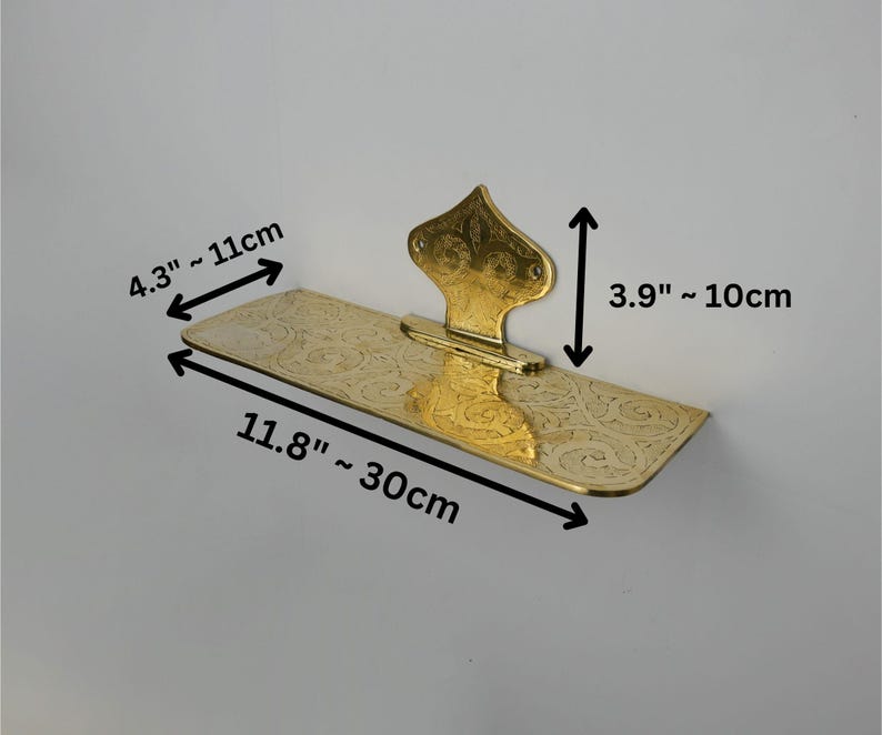 May include: A gold-colored, decorative shelf with an ornate floral design. The shelf is 11.8 inches long, 4.3 inches wide, and 3.9 inches tall. The shelf is mounted on a white wall.