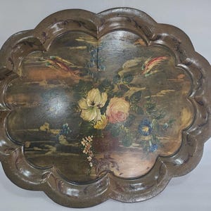 May include: An antique, scalloped-edge serving tray with a painted floral design. The tray features a dark brown border and a central image of flowers in shades of yellow, pink, and blue. The background has a landscape scene with birds.