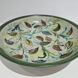 Puede incluir: Un tazón grande y redondo de cerámica con un borde verde y un interior blanco. El tazón está decorado con un diseño pintado a mano de hojas estilizadas en tonos de verde, marrón y amarillo.