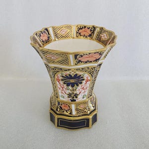 May include: A decorative ceramic vase with a flared top and a hexagonal base. The vase features a detailed floral pattern in navy blue, peach, and gold, with a white interior. The rim has a scalloped edge.