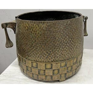 Könnte beinhalten: Ein gehammerter Messing-Pflanztopf mit strukturierter Oberfläche und einem gewebten Muster am Boden. Der Pflanztopf hat zwei verzierte Griffe und einen dunklen Innenraum. Ein dekoratives Stück für den Innen- oder Außenbereich.