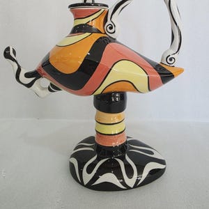 May include: A decorative ceramic lamp with a distinctive design. The lamp has a body with black, orange, and yellow stripes, a white handle with black stripes, and a black and white base. A decorative art piece.