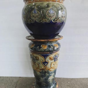 May include: A large, ornate ceramic planter and pedestal set. The planter is a deep blue with green and brown accents, featuring a repeating floral design. The pedestal has a matching design and color scheme, creating a cohesive look. The set is suitable for indoor or outdoor use.