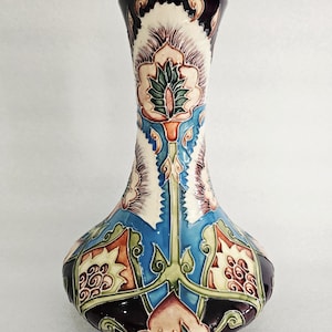 May include: A decorative ceramic vase with a bulbous base and a slender neck. The vase features a detailed floral design in shades of blue, green, orange, and burgundy against a cream background. The vase is approximately 25 cm tall.