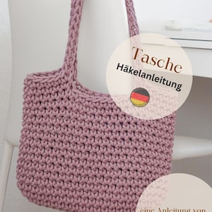 Könnte beinhalten: Eine gehäkelte Tasche in einem staubigen Rosaton. Die Tasche ist mit einem einfachen Stichmuster gearbeitet und hat einen breiten, flachen Boden. Die Tasche hängt an einem weißen Stuhl mit dem Text "Tasche Häkelanleitung" und einer deutschen Flagge.