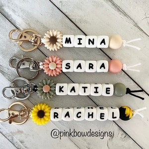 May include: Four personalised keychains with white beads spelling out the names Mina, Sara, Katie, and Rachel. Each keychain features a different flower charm: a white daisy, a pink daisy, a green daisy, and a yellow sunflower.