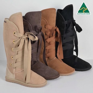 May include: Four tall suede boots in beige, dark brown, tan, and black. Each boot features gold-tone metal eyelets and wide ribbon laces tied in a bow at the front. The boots have a flat sole and a logo in the corner reading Australian Made.