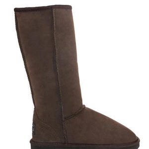 May include: A tall, dark brown suede boot with a rounded toe and a flat, dark brown rubber sole. The boot features visible stitching along the shaft and a small, rectangular black brand label located on the lower outer side near the heel area.