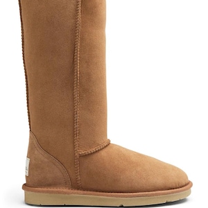 May include: A tall, tan-coloured suede boot with a rounded toe and a flat, light-coloured rubber sole. The boot features visible vertical stitching along the shaft and a small, rectangular white brand label on the lower heel area.