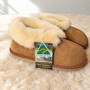May include: Tan suede slippers with a thick, cream-colored sheepskin lining and a flat, light brown sole. A rectangular tag is attached to the front, featuring a green kangaroo logo, the text AUSTRALIAN MADE, and 100% Sheepskin.
