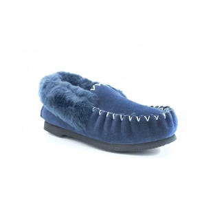 May include: A navy blue suede moccasin slipper featuring a thick, plush navy blue faux fur lining around the collar. The upper is accented with white whipstitch detailing along the toe seam and a black rubber sole for traction and support.
