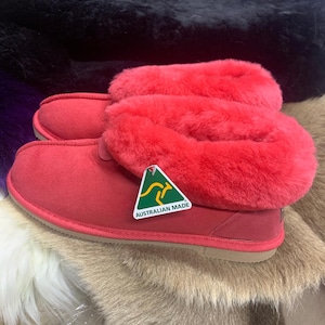May include: A pair of bright red suede slippers featuring a thick, plush red faux fur lining and a tan rubber sole. A triangular green and gold tag attached to the side displays a kangaroo graphic and the text AUSTRALIAN MADE.