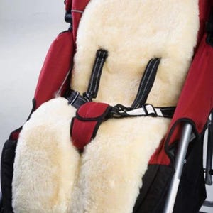 May include: A cream-colored, plush sheepskin stroller liner featuring cutouts for a five-point safety harness. The thick, soft fleece insert is positioned inside a red and black stroller, providing a cushioned seat and backrest surface.