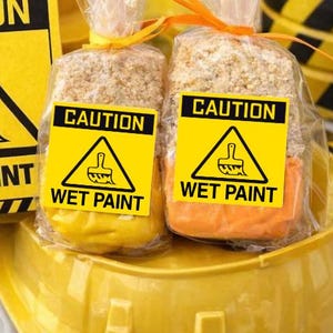 May include: A construction-themed birthday party setup with yellow and black decorations. Includes a toy construction truck, a sign that reads "CAUTION WET PAINT", and packaged treats with the same warning. The theme is reinforced with a yellow hard hat.